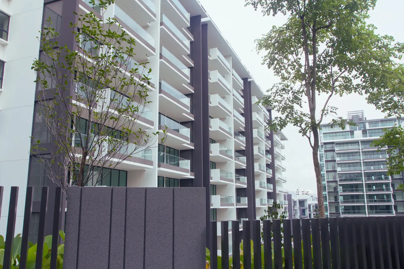 Modern apartment buildings with white balconies and dark accents, surrounded by trees and a dark fence in the foreground.