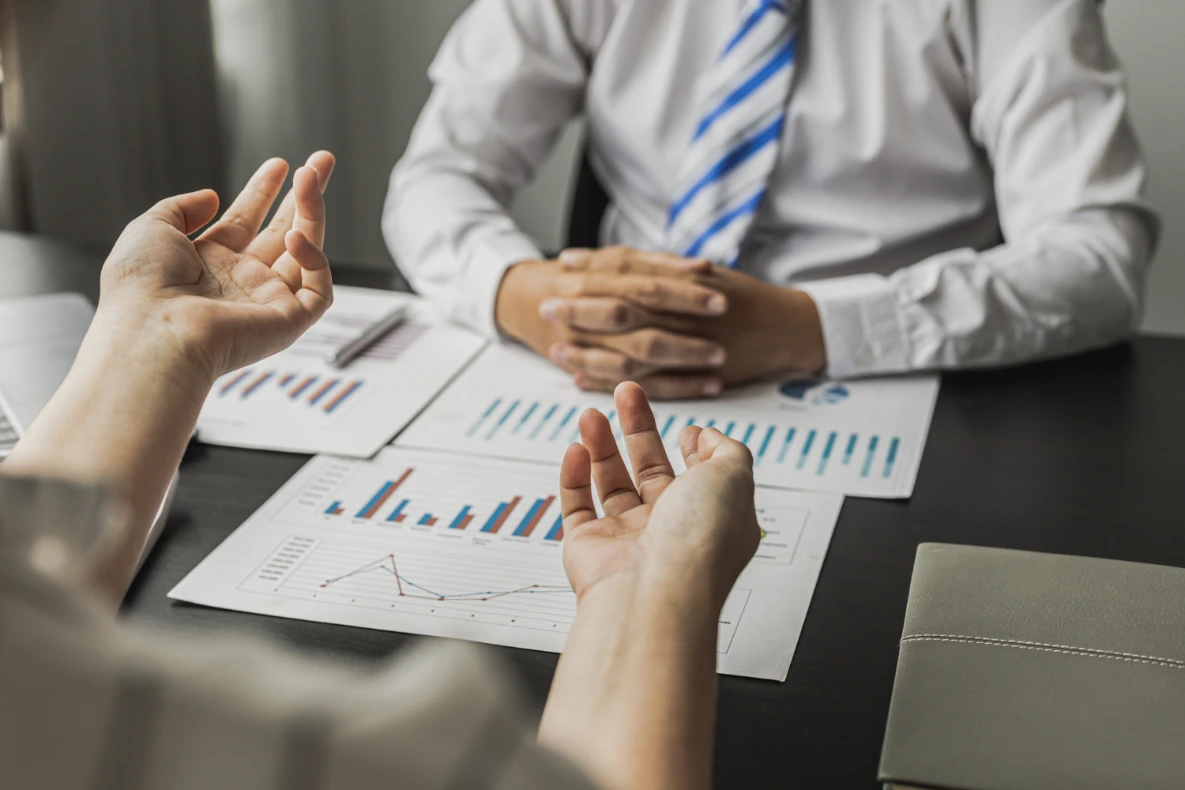 Two individuals are seated at a desk, reviewing financial charts and graphs. One person's hands are gesturing while speaking, and the other person has their hands clasped in front of them. The scene suggests a business meeting or discussion.