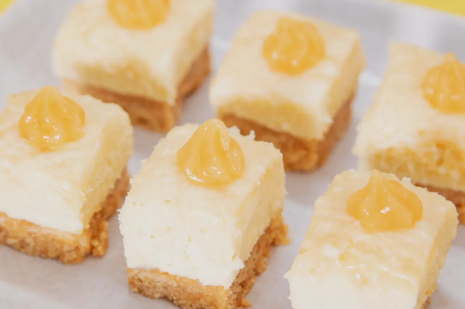 A close-up of six squares of cheesecake with a yellow fruit topping on a light-colored plate.