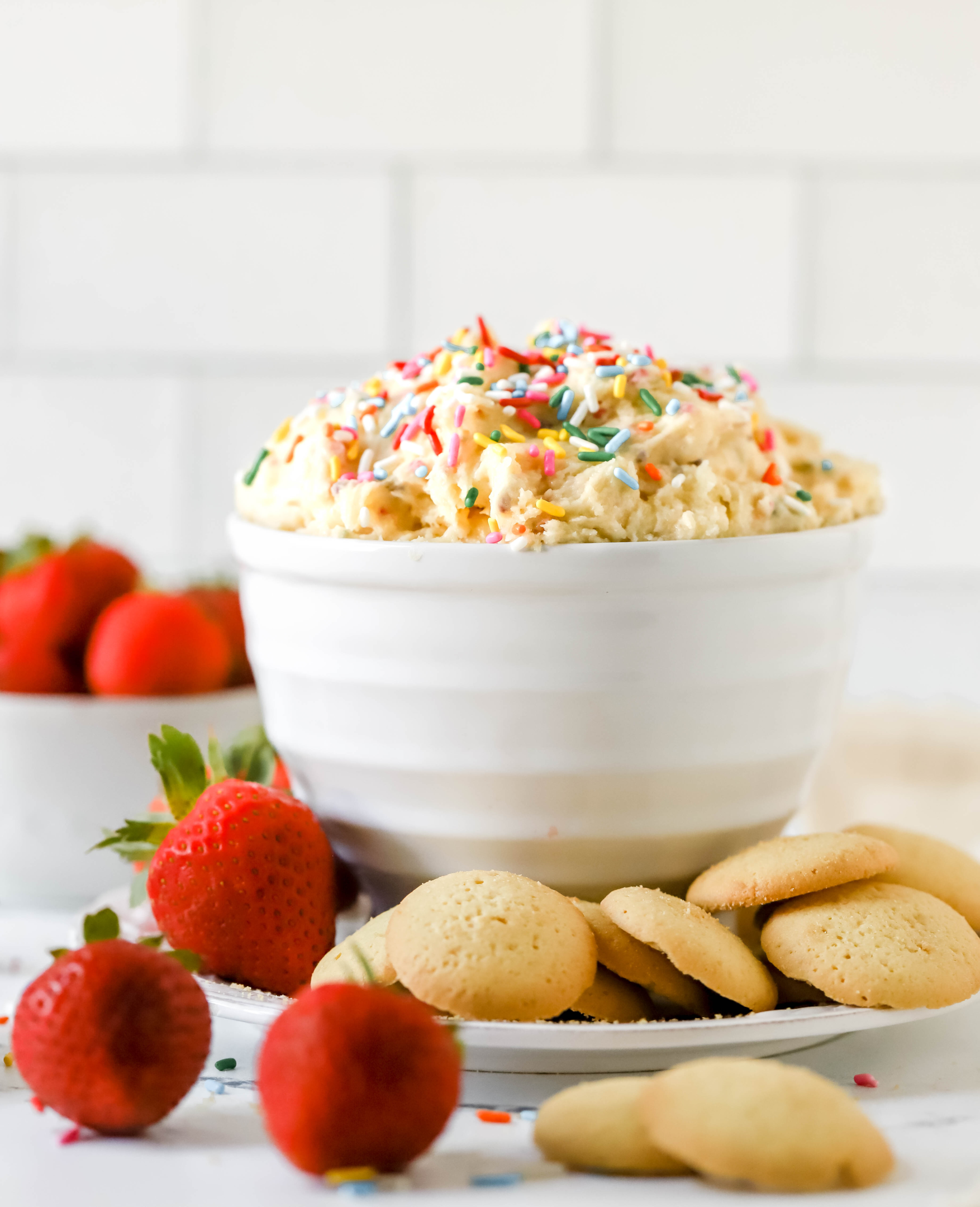 side view of a bowl of cake batter dip in a bowl on a plate with strawberries and cookies