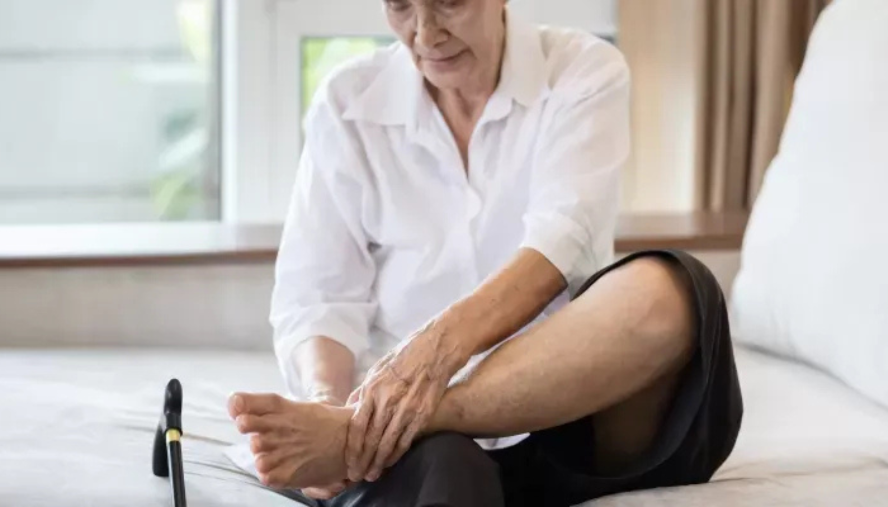 Eldre person sitter på seng og masserer venstre ankel; smerter i foten ved nevropati.