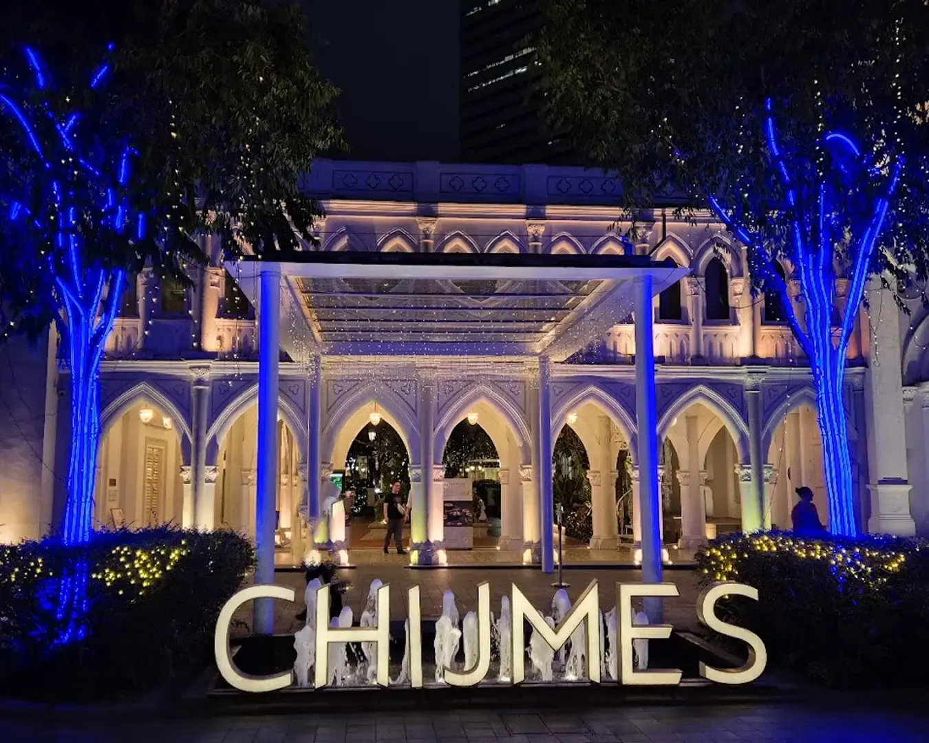 Illuminated arches of a historic building are adorned with blue and white lights, with "CHIJMES" sign in front, creating a festive nighttime ambiance.