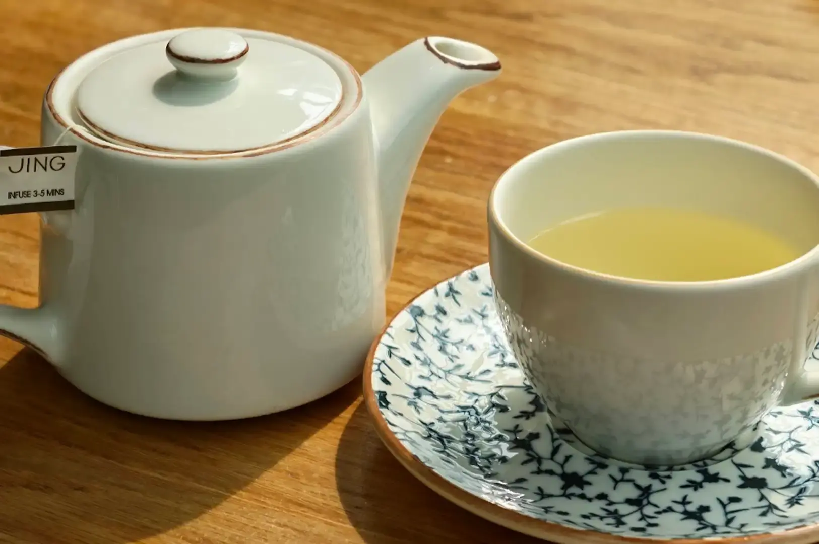 A bright close-up shot on a light wood surface featuring a white ceramic teapot with a JING tea tag resting against its handle, situated next to a teacup filled with clear pale-yellow tea resting on a blue-and-white floral patterned saucer.