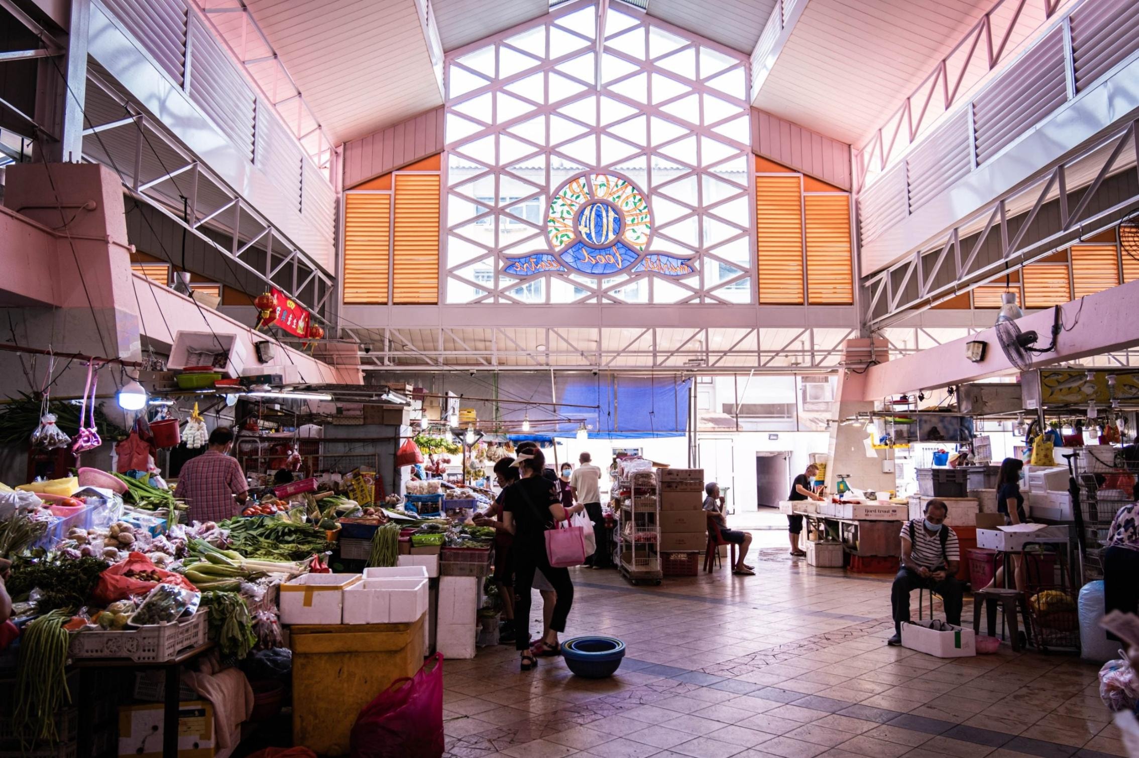  This interior view showcases a bustling wet market filled with fresh produce stalls and shoppers under a high, vaulted ceiling. Natural light streams through a large geometric window feature that prominently displays the "Chong Pang Market & Food Centre" logo.