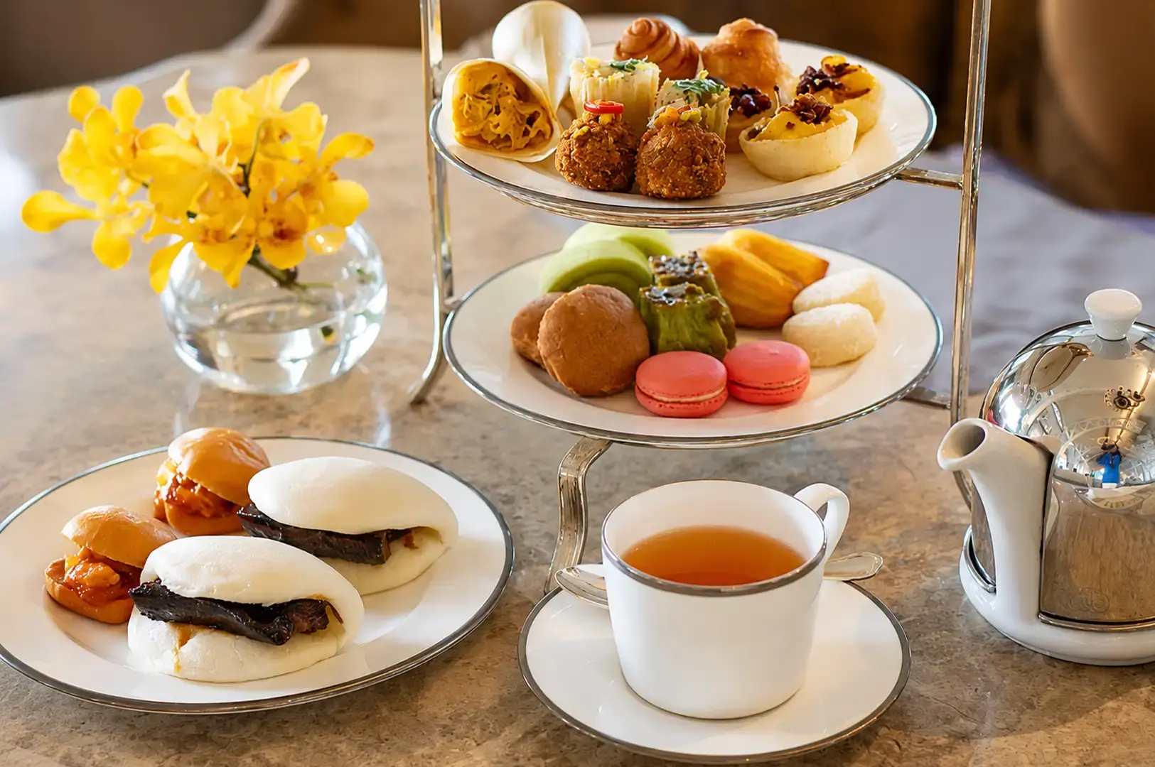 An afternoon high tea spread centered around a three-tier silver stand loaded with sweet pastries, macarons, and savory bites, accompanied by a plate of fluffy white bao buns filled with dark braised meat, a teacup filled with warm tea, a shiny metallic teapot, and a small glass vase of bright yellow orchids.