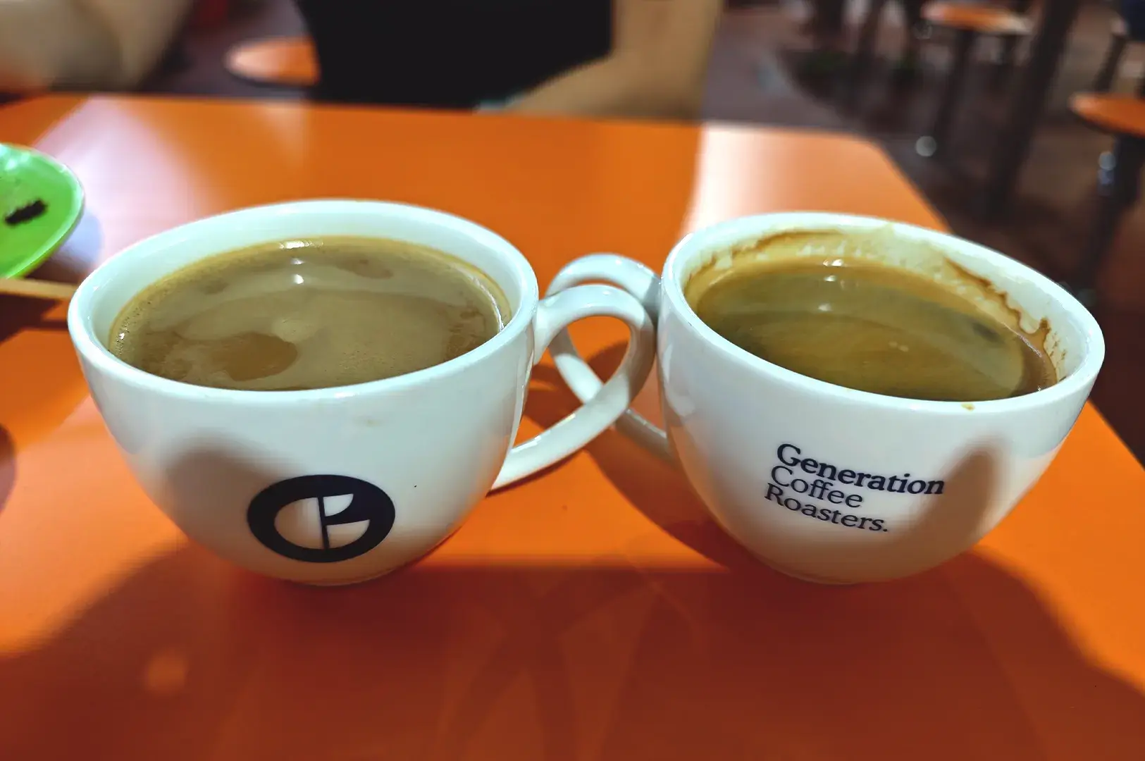Two hot coffee cups of single origin and specialty coffee placed side by side on an orange table, showcasing the branded mugs from Generation Coffee Roasters.
