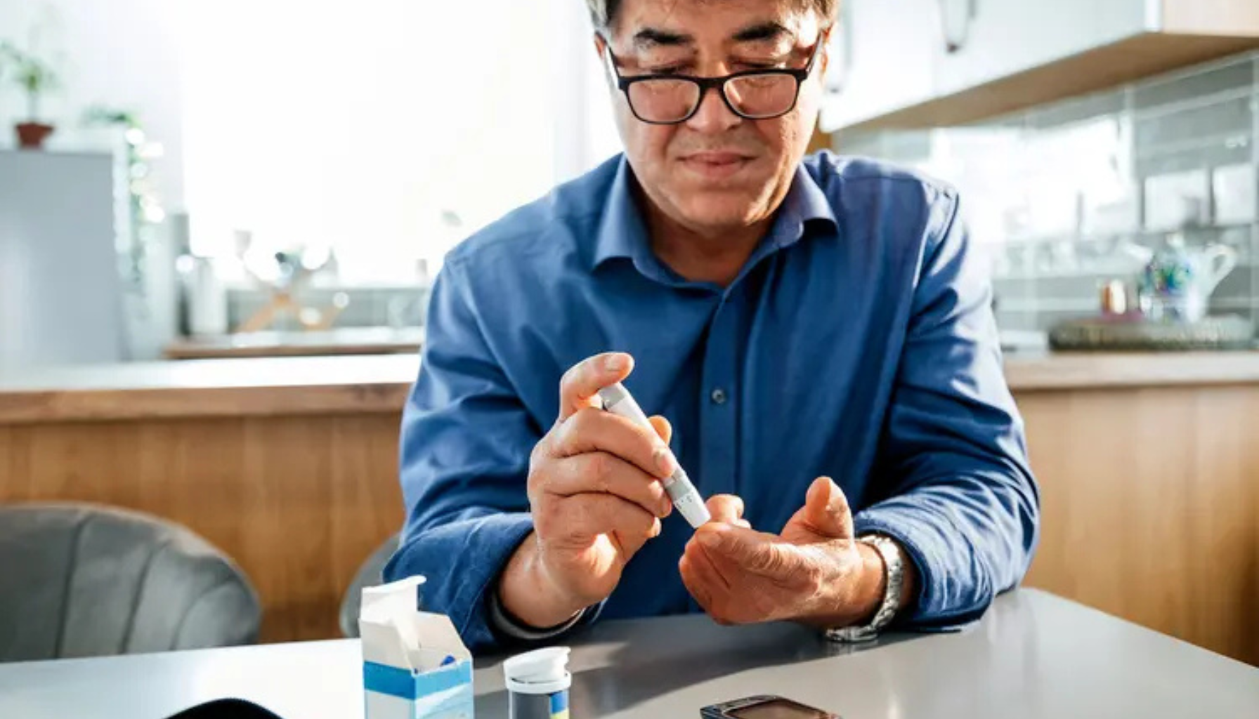 Mann i blå skjorte måler blodsukkeret sitt hjemme ved hjelp av fingerprikktest og blodsukkerapparat på kjøkkenbordet.