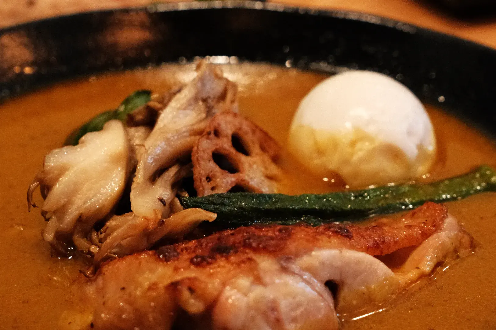 A plate of food featuring a piece of grilled chicken, lotus root, green beans, sautéed mushrooms, and a poached egg in a rich sauce.