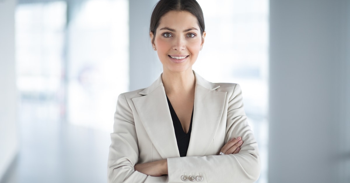 Confident woman in a light blazer with arms crossed, standing in a bright, modern office, offering tax solutions.