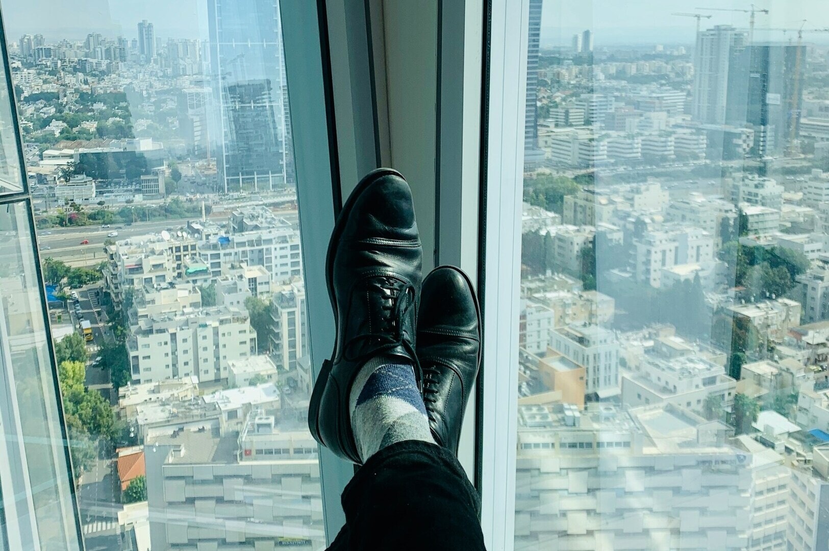 A person in black shoes and patterned socks relaxes with feet up, overlooking a cityscape through a tall window; conveys a sense of relaxation and leisure.