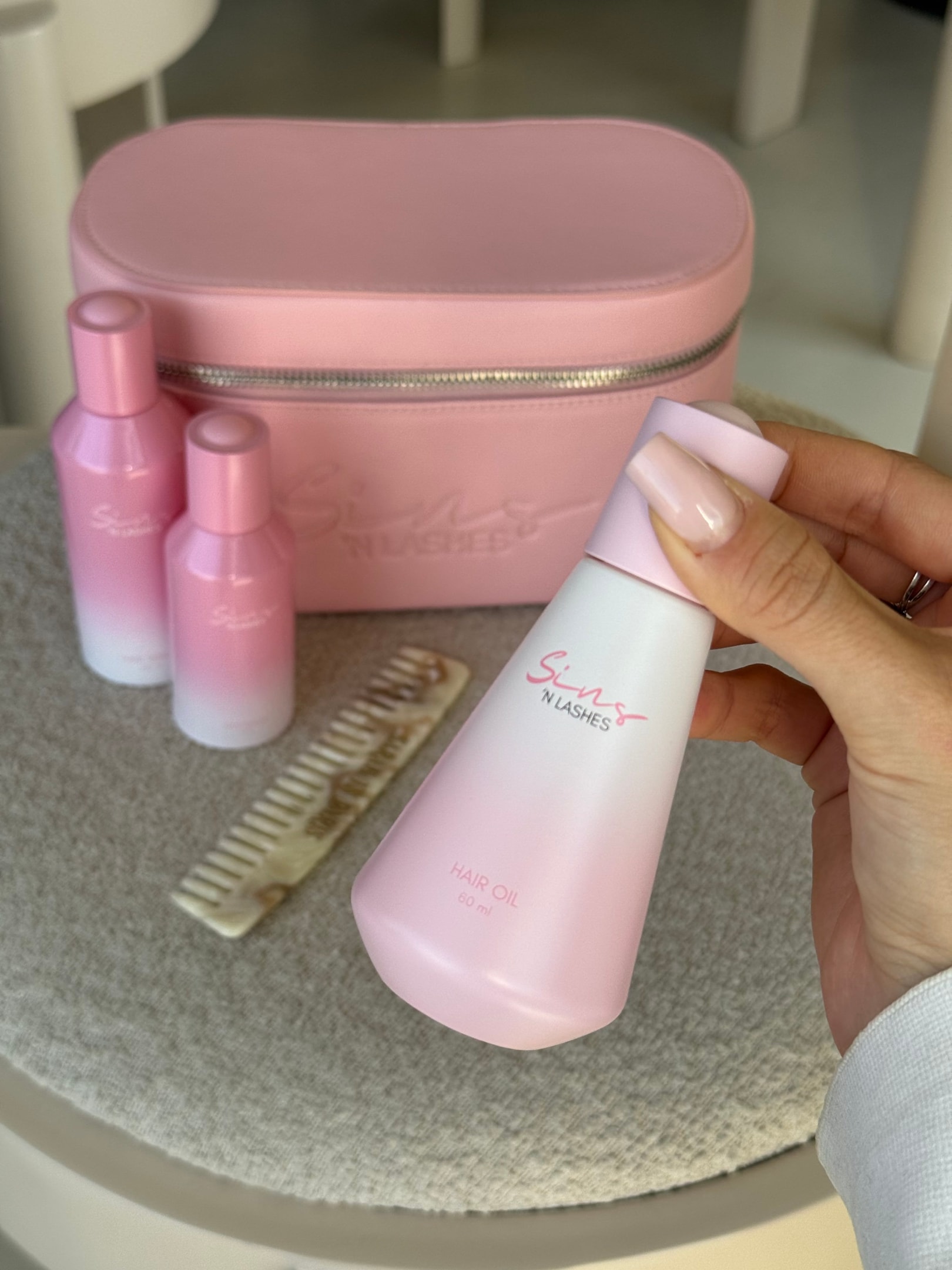 Hand hält die rosa Sins ’N Lashes Hair-Oil-Flasche; im Hintergrund stehen passende Haircare-Produkte, ein Kamm und eine rosa Beauty-Bag – perfekte Haarpflege-Routine für gesundes, glänzendes Haar.