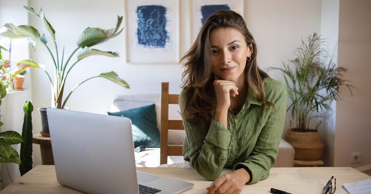 Smiling woman at desk with plants and laptop, thinking about OnlyFans consumption tax vs income tax.