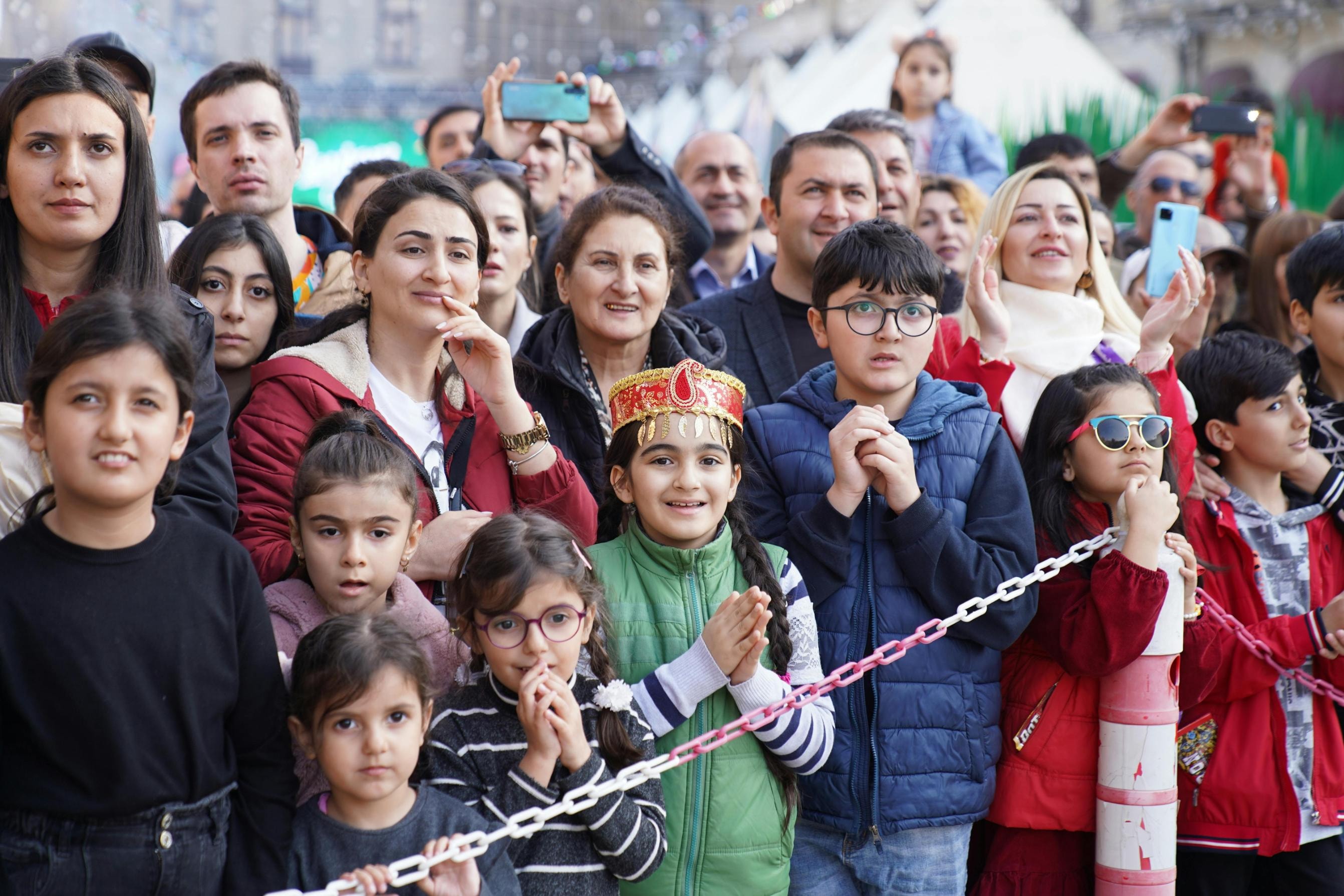 This image captures a large, attentive crowd of adults and children gathered outdoors, appearing engaged as they look forward from behind a low chain barrier. A young girl in the foreground wearing a decorative red and gold headdress serves as a focal point, while several people in the background hold up smartphones to record the event.