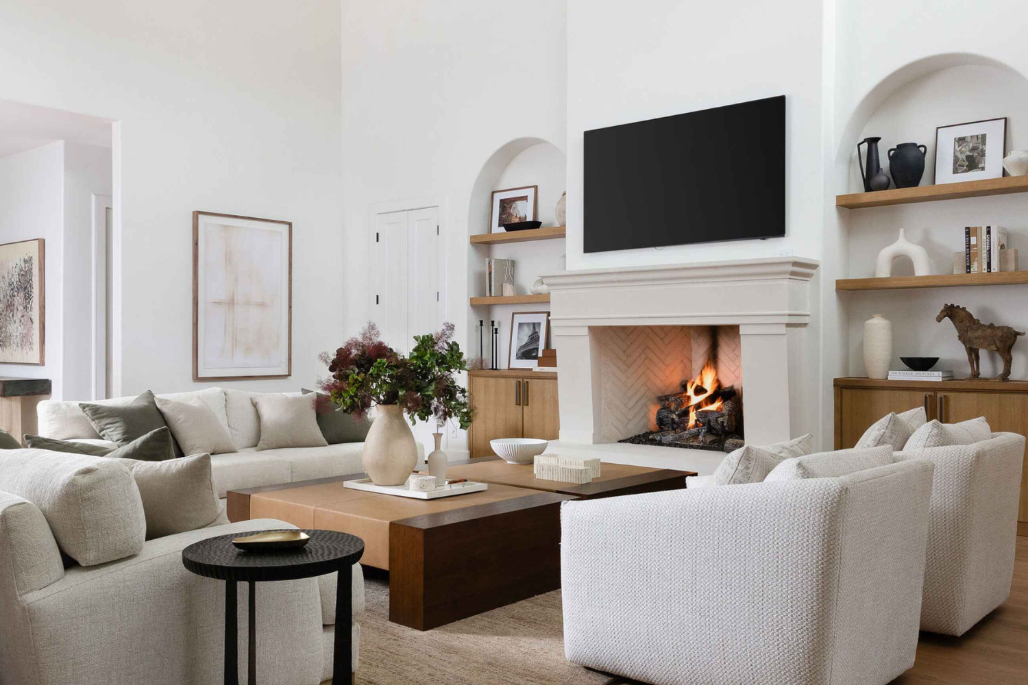 Warm and welcoming living room with layered white sofas, a central fireplace, natural wood elements, and built-in shelving styled with neutral decor.