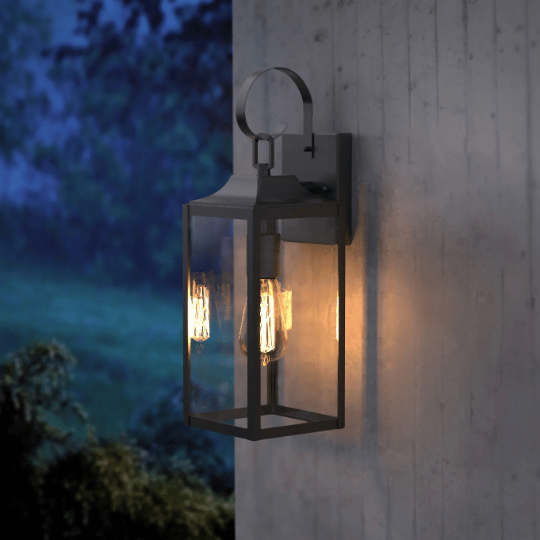 Modern black outdoor wall lantern with clear glass panels and glowing filament bulb, mounted on a concrete wall at dusk.